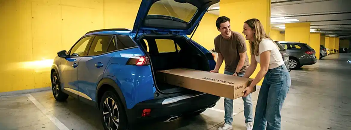 IKEA alışverişi sonrası büyük mobilya kutusunu otoparkta mavi Peugeot SUV bagajına sığdıran mutlu çift ve hacimli yükler için günlük araç kiralama çözümü.